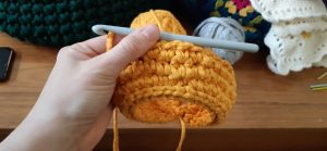 Up close view of Kelly's orange crochet sample, held with the crochet needle inserted and ballf of yarn and crochet items in the background