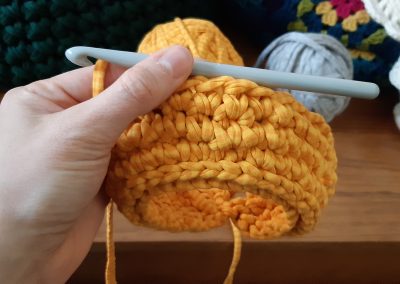 Up close view of Kelly's orange crochet sample, held with the crochet needle inserted and ballf of yarn and crochet items in the background