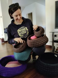 Kelly smiling looking at many crochet products balancing on her knees and on the wooden floor in front of her
