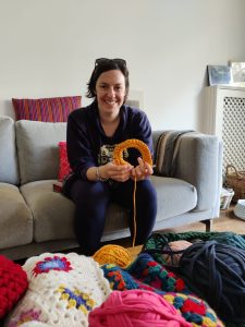Kelly holding a crochet sample sat on a sofa with yarn and crochet in front at an IWAM Creatives event