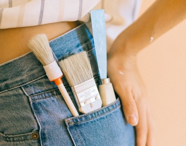 Close up picture of a woman wearing jeans showing her back pocket with paint brushes and a scraper ready for DIY work