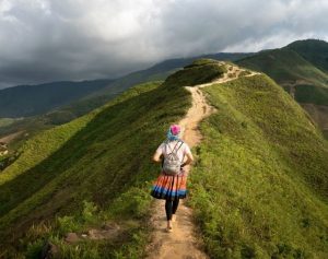 Woman walking away alone, along a ridge on top of a steap hill wearing a skirt, leggings, t shirt, backpack and hat