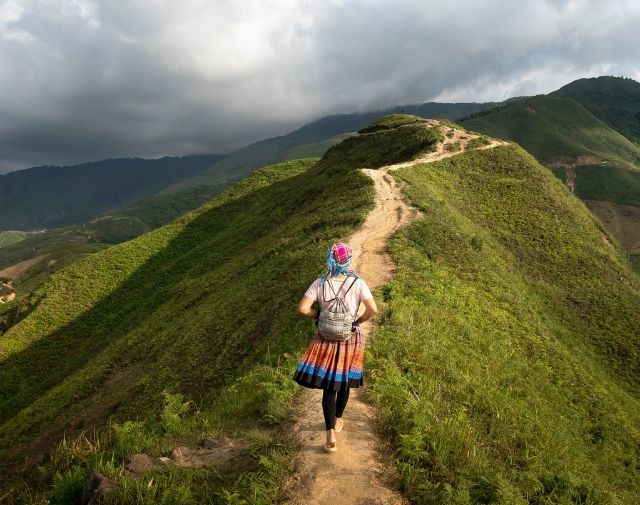 IWA Modena Walking Hiking Interest Group Woman walking away alone, along a ridge on top of a steap hill wearing a skirt, leggings, t shirt, backpack and hat
